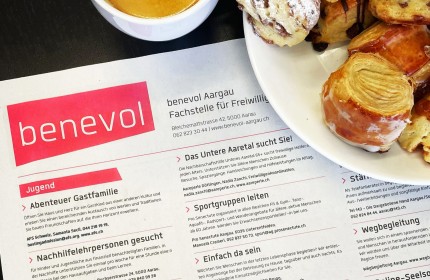 Zu sehen ist ein Foto des Freiwilligen-Stellenanzeigers von benevol Aargau in einer Zeitung sowie Teile eines Kaffees und einem Teller mit Kaffeegeb&auml;ck.